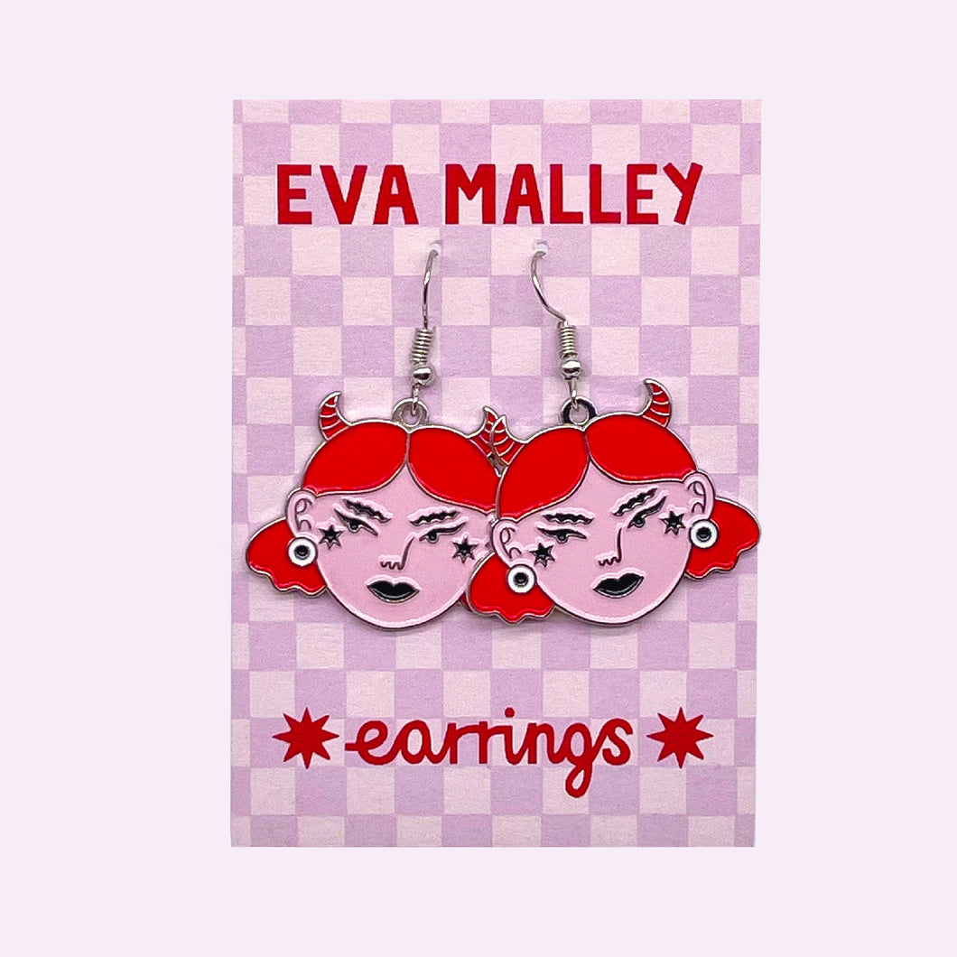 A pair of earrings, girls with an angry expression, pale pink faces, bright red hair and horns. They are attached to a lilac checkerboard earring backing card.